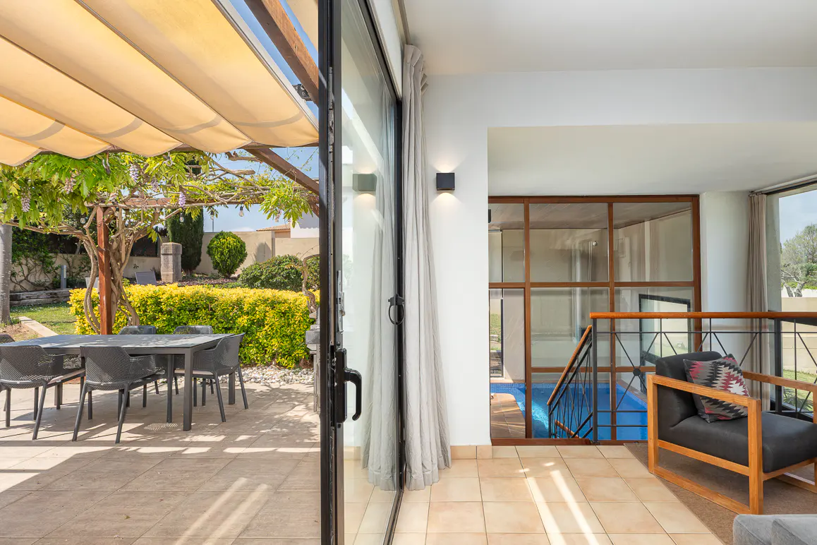Indoor-outdoor living space with patio, dining set, and pool view through glass wall. Neutral tones and natural light.
