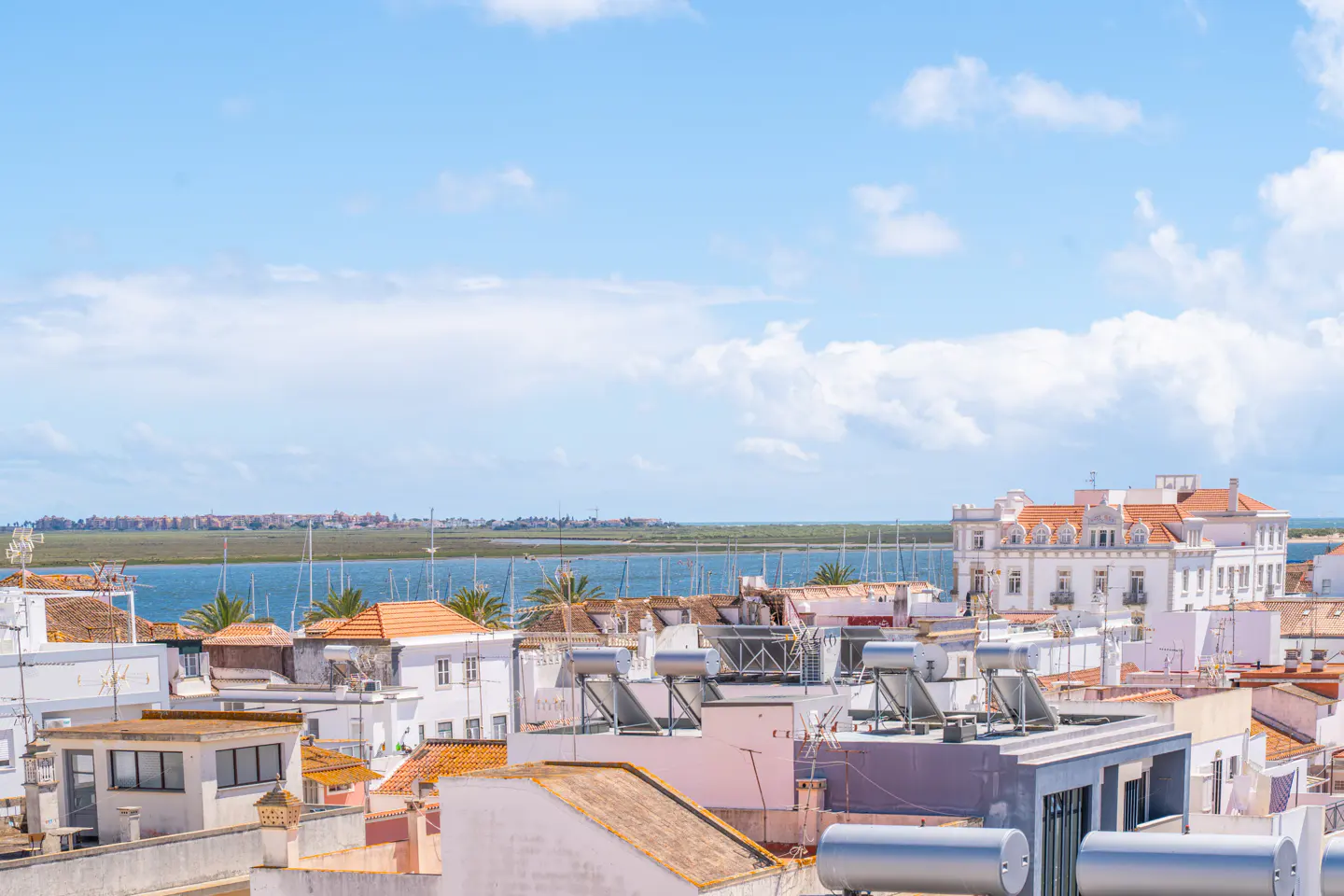 Rooftop view of Tavira, Portugal, with white buildings, orange tile roofs, and solar panels. Blue water and sky in the background.