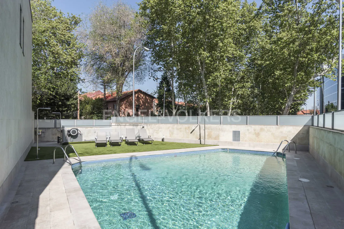 Outdoor pool with clear blue water, surrounded by lounge chairs, green grass, and trees. Building in background.