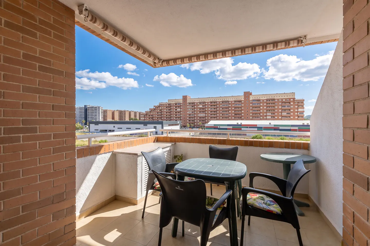 Balcony with brick walls, a table and chairs, and a view of buildings under a blue sky with clouds.