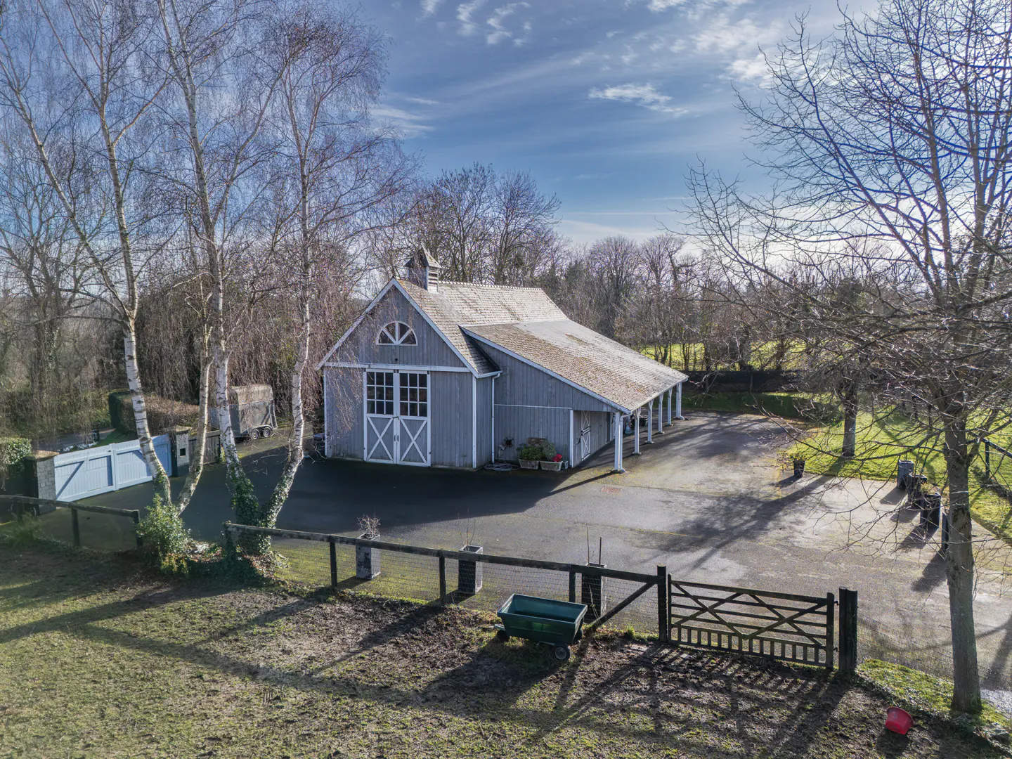 A gray barn with white trim sits on a paved lot, surrounded by trees and a wooden fence.