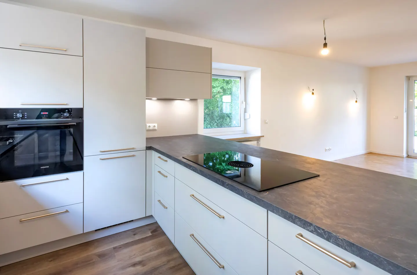 Bright kitchen with white cabinets, gold handles, and a dark countertop island with a black cooktop. A window and lights illuminate the space.