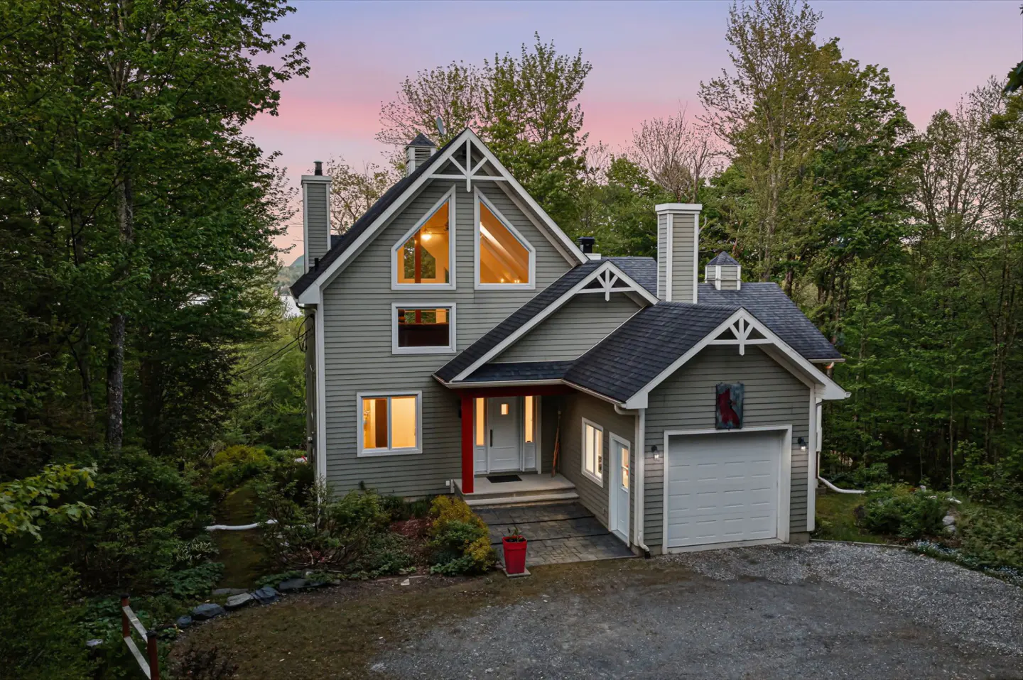A two-story grey home with a white garage and red accent pillar, surrounded by lush trees at sunset.