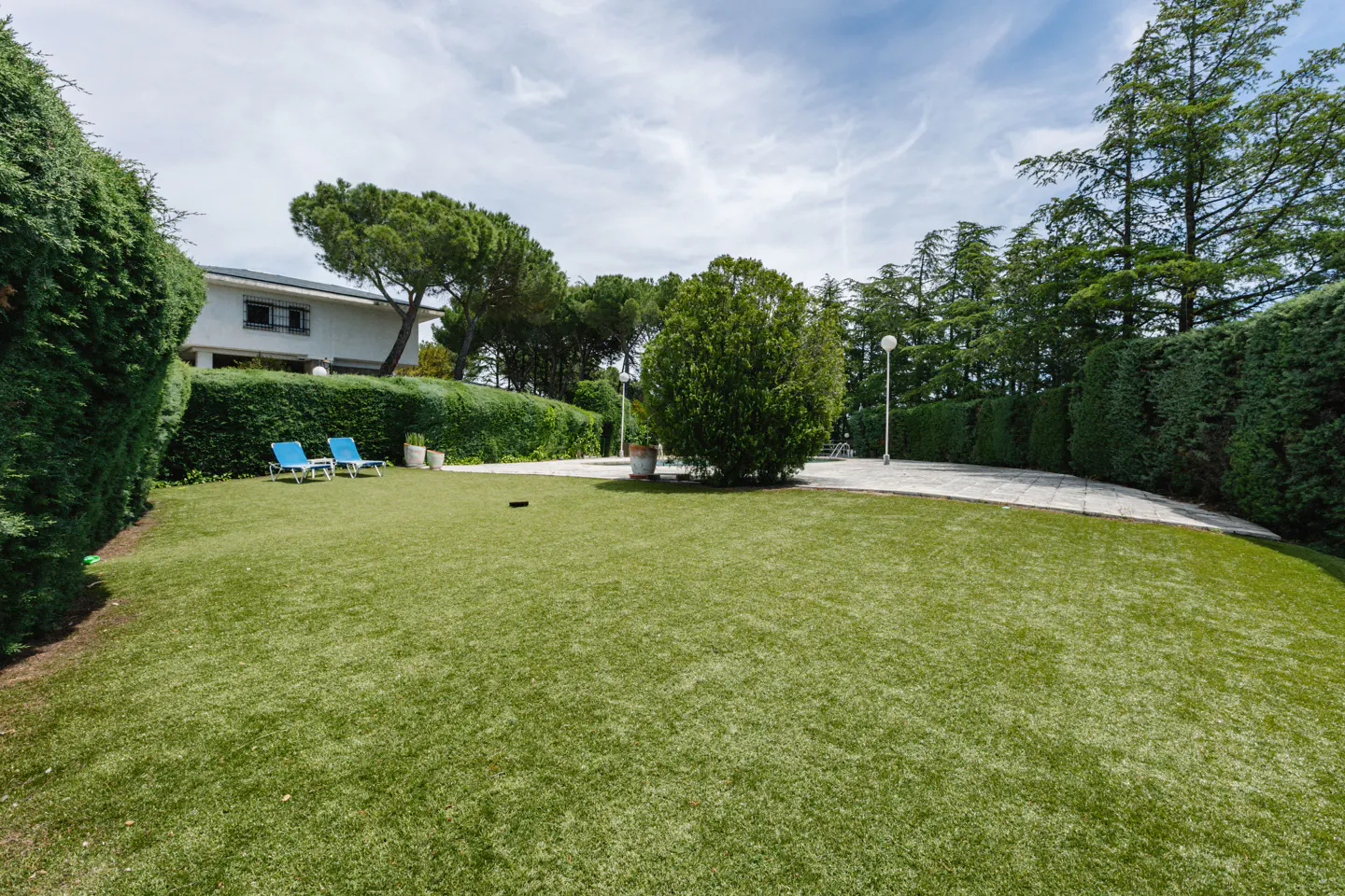 A wide shot of a green lawn with two blue lounge chairs, hedges, trees, and a house in the background.