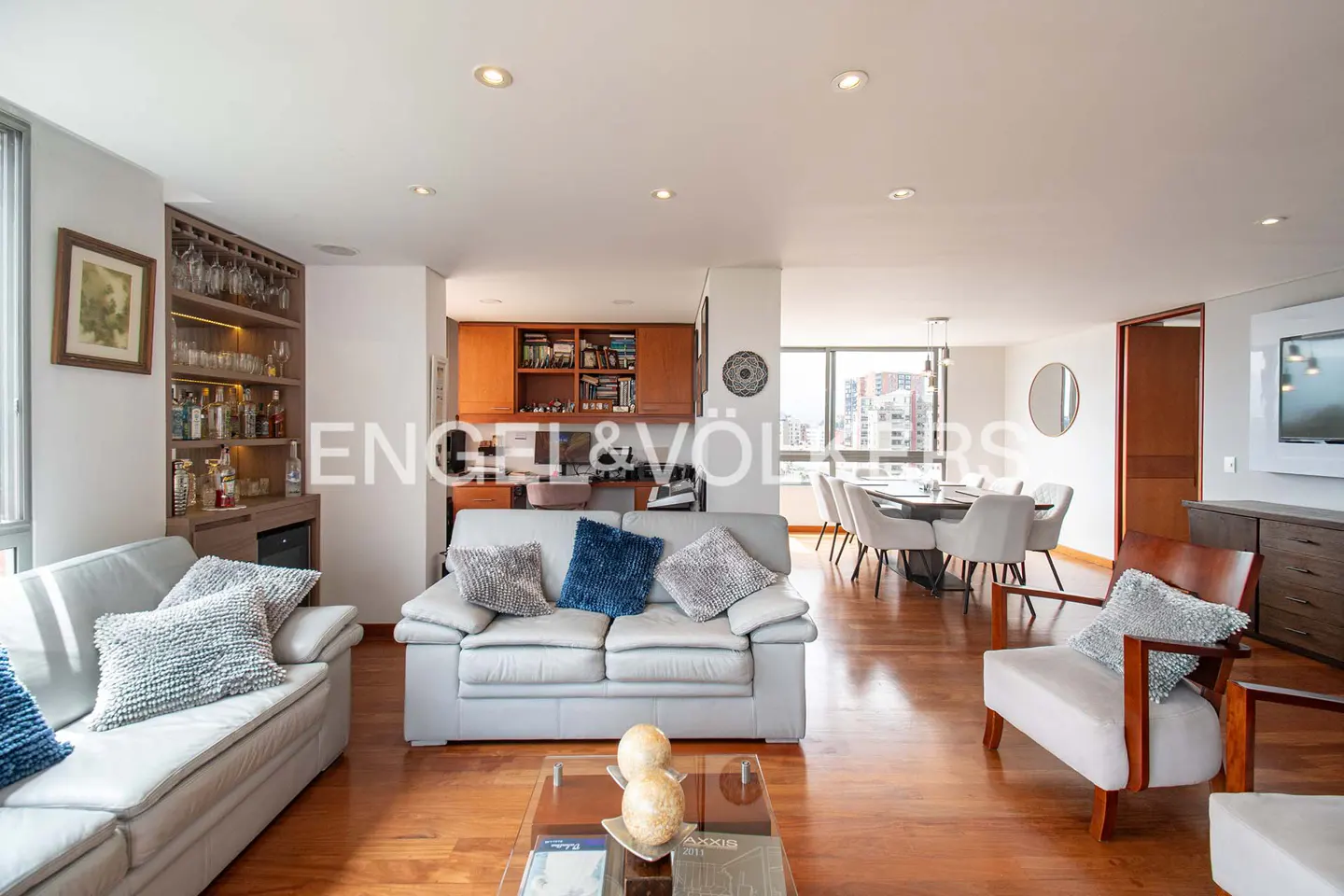 Living room with light gray sofas, wood floors, and a dining area with city views. Built-in bar and desk area visible.