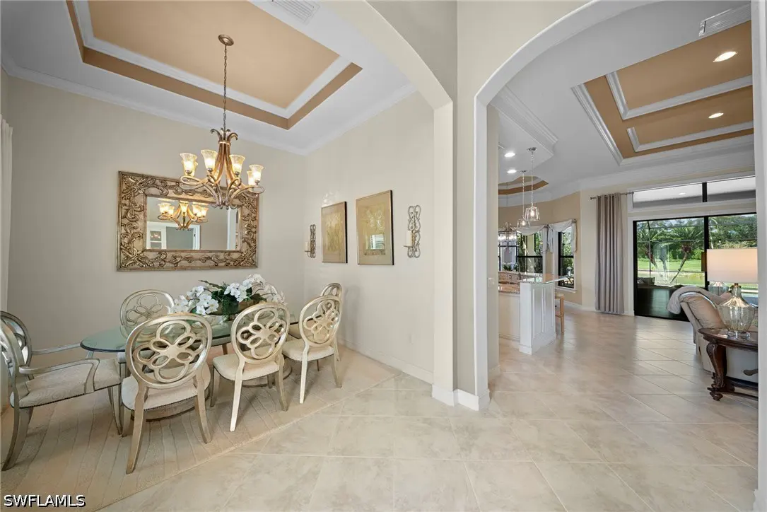 A dining room with a glass table, ornate chairs, and a gold chandelier. The room has beige walls and tile floors, with an open view to the kitchen and living room.