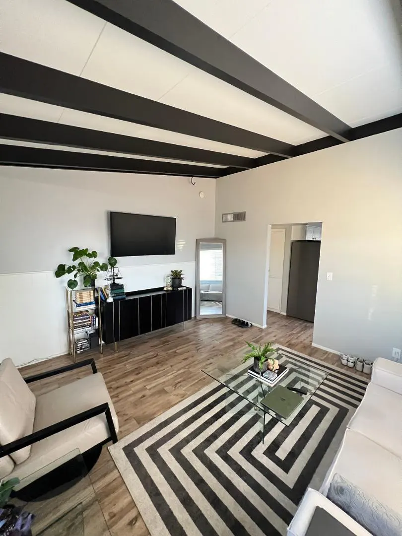 Living room with wood floors, black ceiling beams, and a black and white geometric rug. A black media console sits under a flat screen TV.