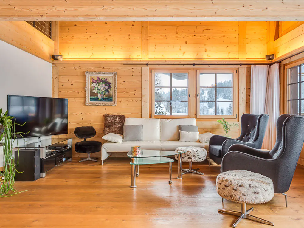 Cozy wooden living room with a white sofa, two dark armchairs, glass coffee tables, and a view of snowy mountains outside.