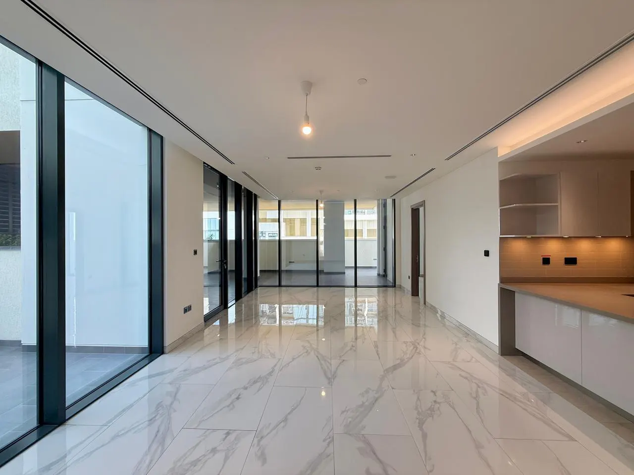 Bright, empty apartment with white marble floors and floor-to-ceiling windows. A modern kitchen is visible on the right.