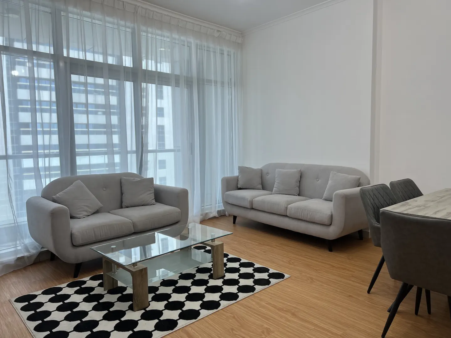 Living room with two gray sofas, a glass coffee table on a black and white rug, and a dining table with gray chairs. Large windows with sheer white curtains.