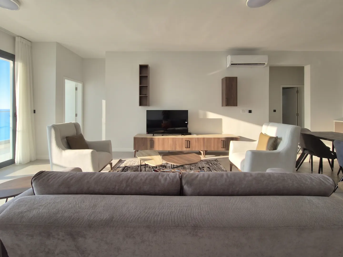 Living room with a gray sofa, two white armchairs, a TV, and a view of the ocean through a glass door.