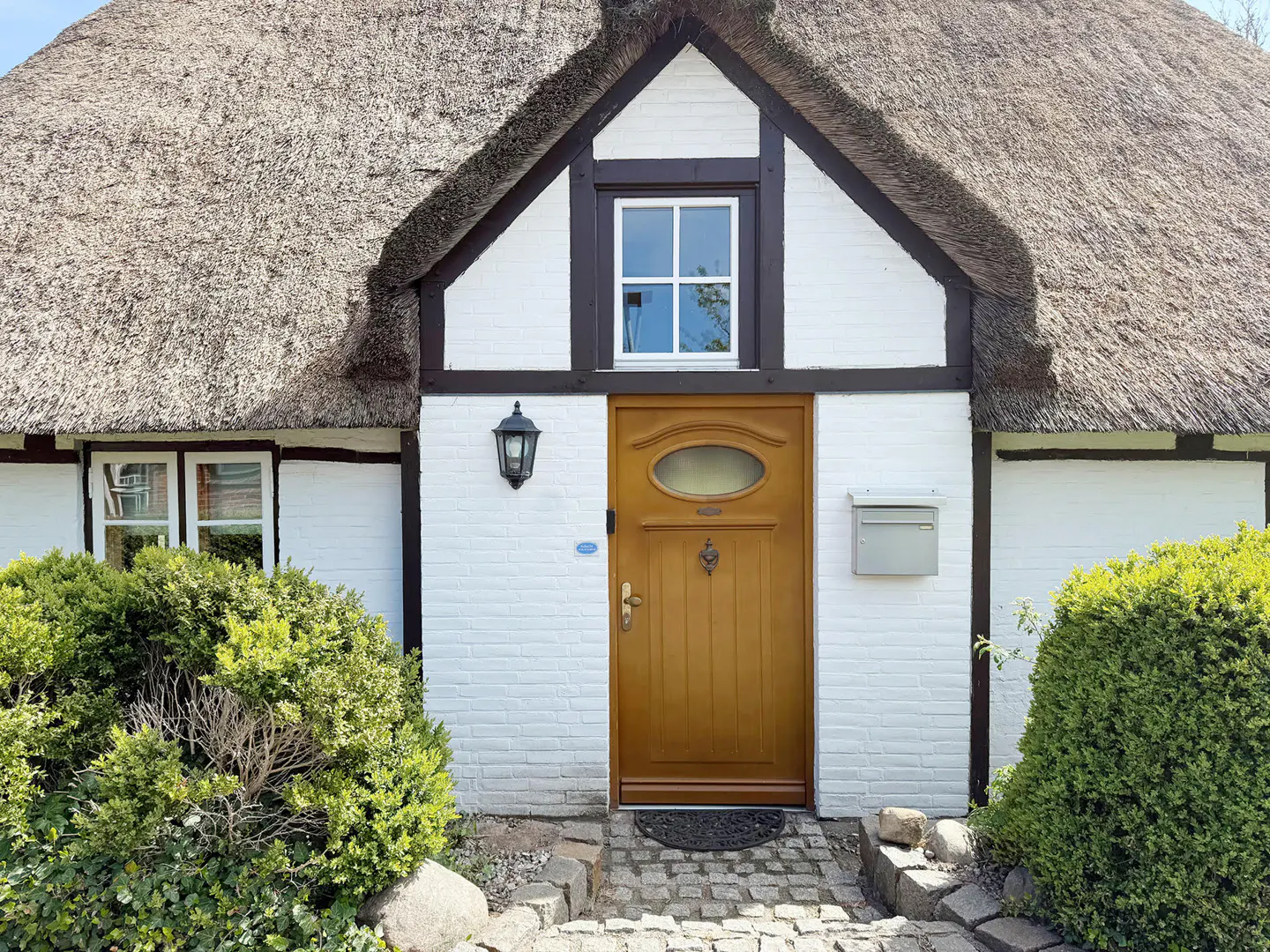A charming white cottage with a thatched roof, a wooden front door, and lush green bushes in the front garden.