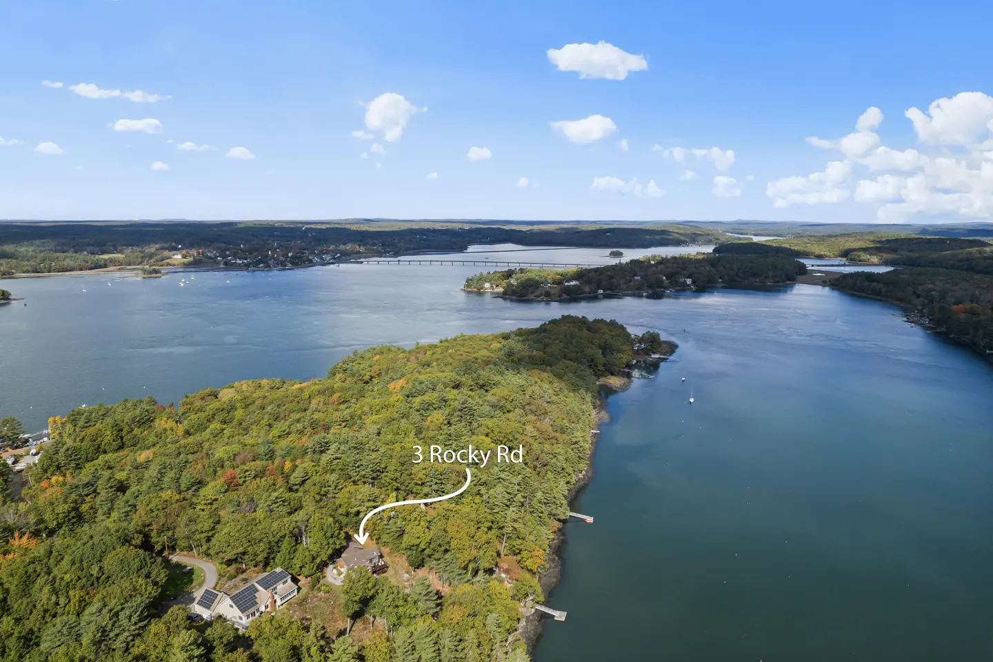 Aerial view of 3 Rocky Rd, a house on a wooded peninsula surrounded by water under a blue sky with scattered clouds.