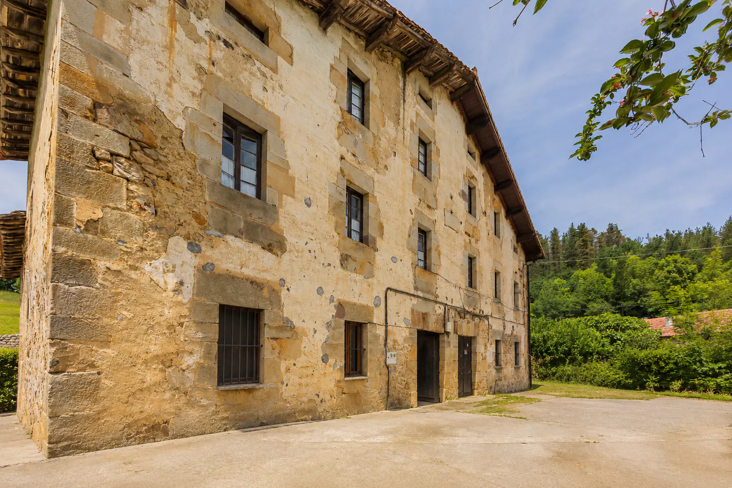 Exterior view of a weathered, multi-story stone building with dark-framed windows and doors, set against a backdrop of lush green trees and a blue sky.