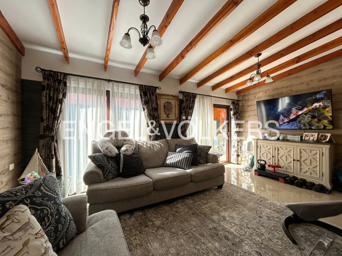 Living room with wood paneled walls, exposed beams, and a beige sofa with decorative pillows. A large TV sits above a white cabinet.