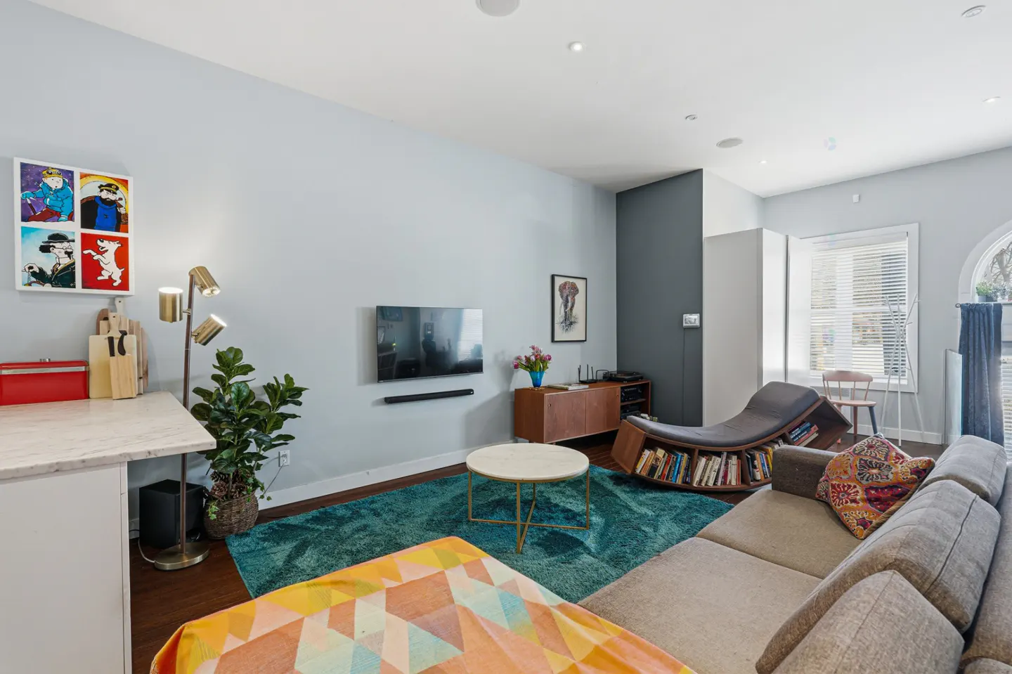 Living room with gray sofa, teal rug, and unique bookshelf chair. TV on the wall, and colorful art adds a modern touch.
