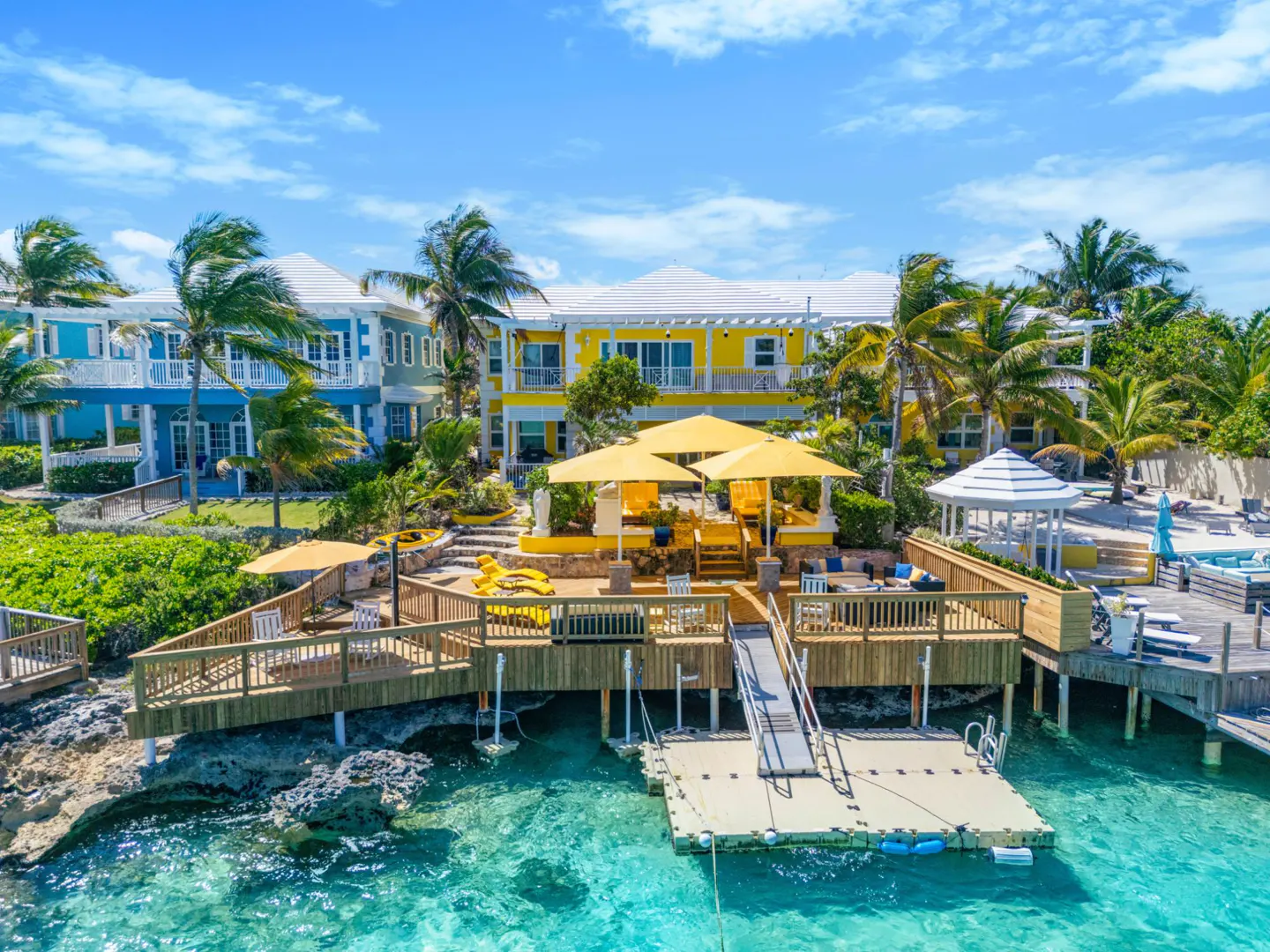 Waterfront property with a yellow house, wooden deck, and dock on turquoise water. Palm trees and blue sky in the background.