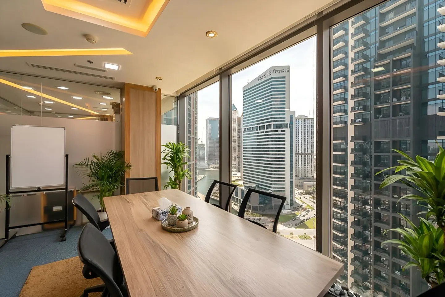 Bright office with a long wooden table, black chairs, and a whiteboard. Large windows offer a city view with skyscrapers.