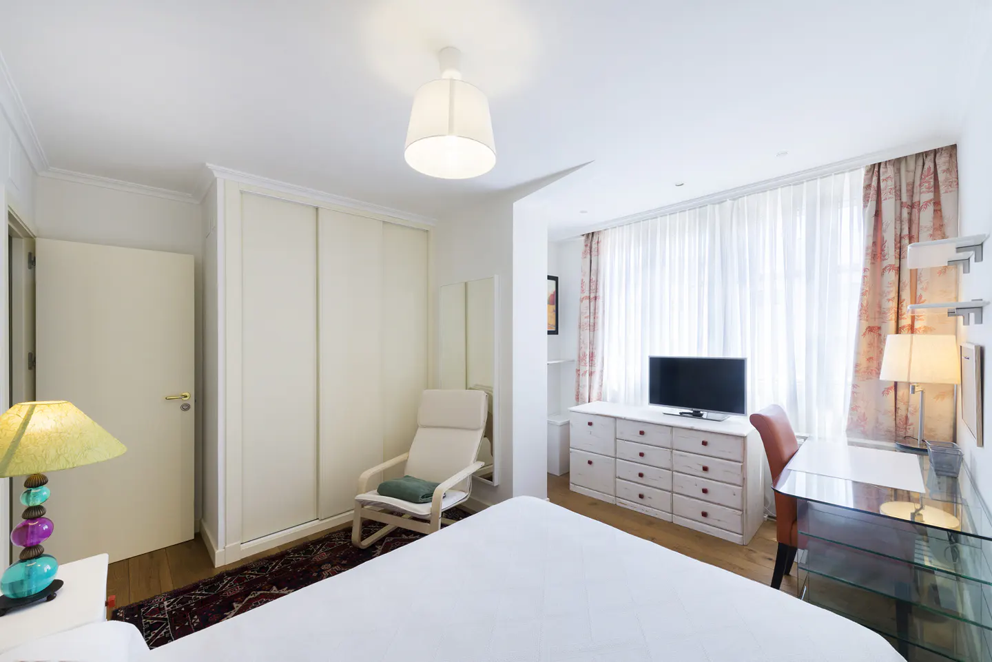 Bright bedroom with white walls, a white bed, a desk, and a TV on a white dresser. A chair sits near a closet.