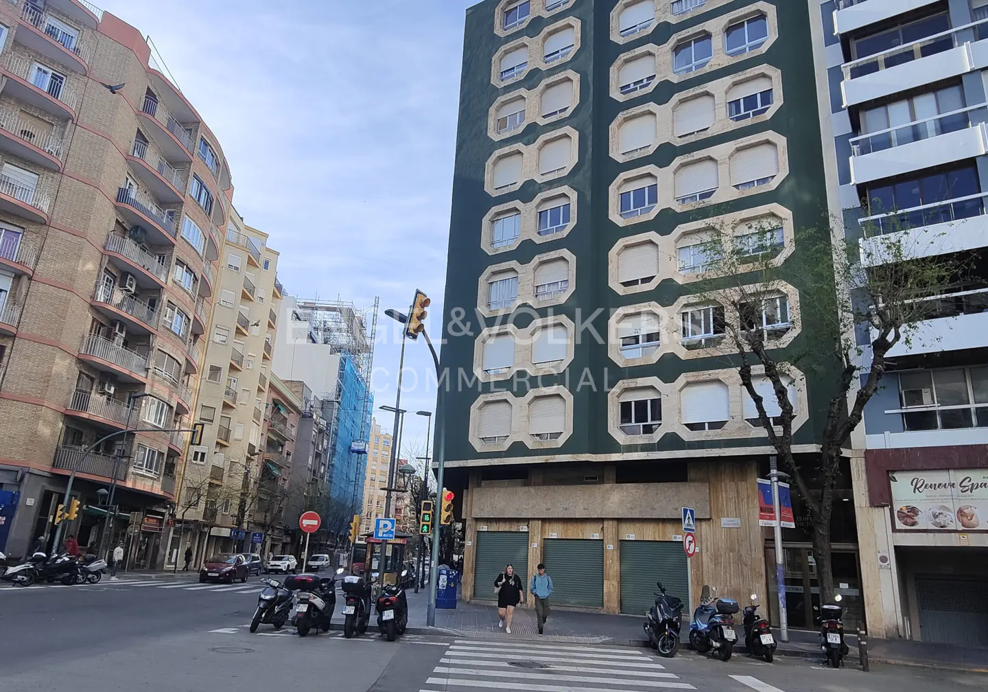 Street view of a tall green building with white window frames, people crossing the street, and parked motorcycles.