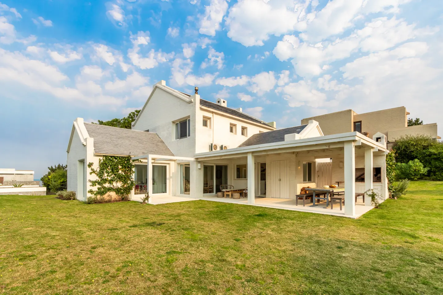 White two-story house with a covered patio, outdoor dining area, and green lawn under a blue sky with scattered clouds.