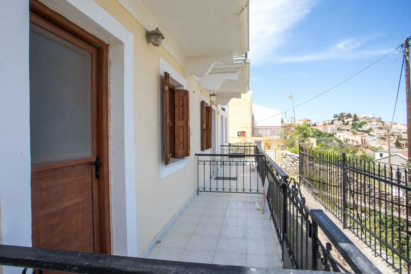 A balcony with a tiled floor, wooden doors, and black metal railings overlooking a hillside town under a clear blue sky.