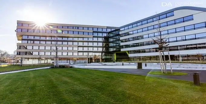 Modern office building with many windows, surrounded by green lawn and trees under a clear blue sky.