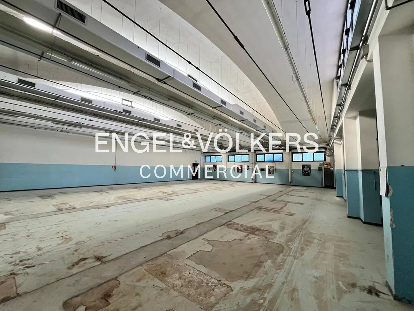 Large, empty commercial space with concrete floors, blue and white walls, and high ceilings with industrial lighting. Engel & Völkers logo overlay.