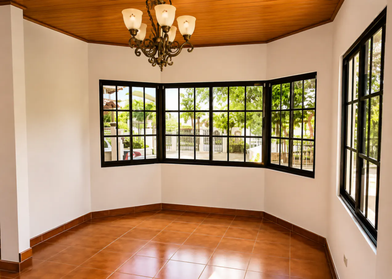 Empty room with brown tile floor, white walls, and wood ceiling. Black framed windows overlook green trees. A chandelier hangs from the ceiling.