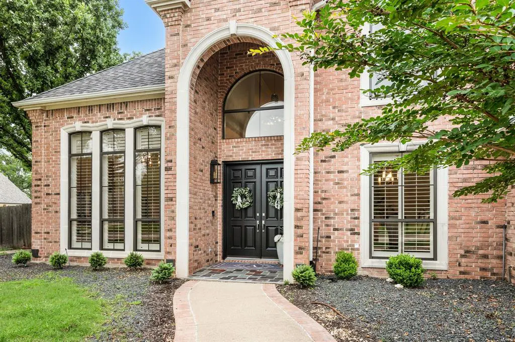 Exterior of a two-story brick house with black double doors and a curved archway. Wreaths hang on the doors. Windows have black frames.