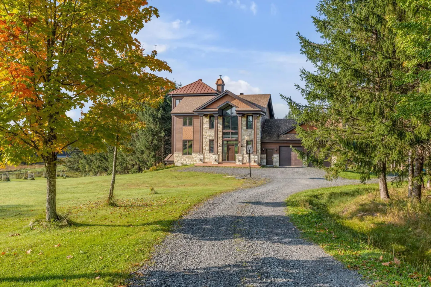 A stone and brown siding house with a gravel driveway, surrounded by green lawns and autumn trees under a blue sky.