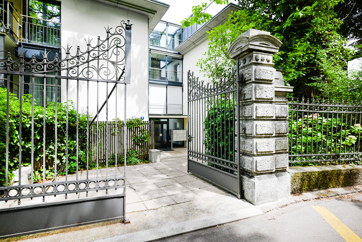 Open wrought iron gates lead to a modern white apartment building entrance surrounded by lush green foliage.
