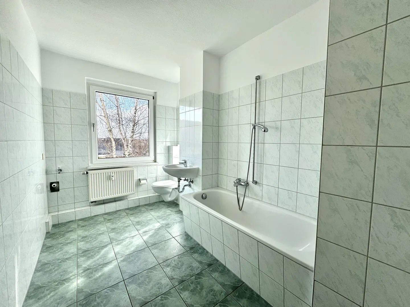 Bright bathroom with green tiled floor, white tiled walls, a white bathtub, toilet, sink, and a window with a view of trees.