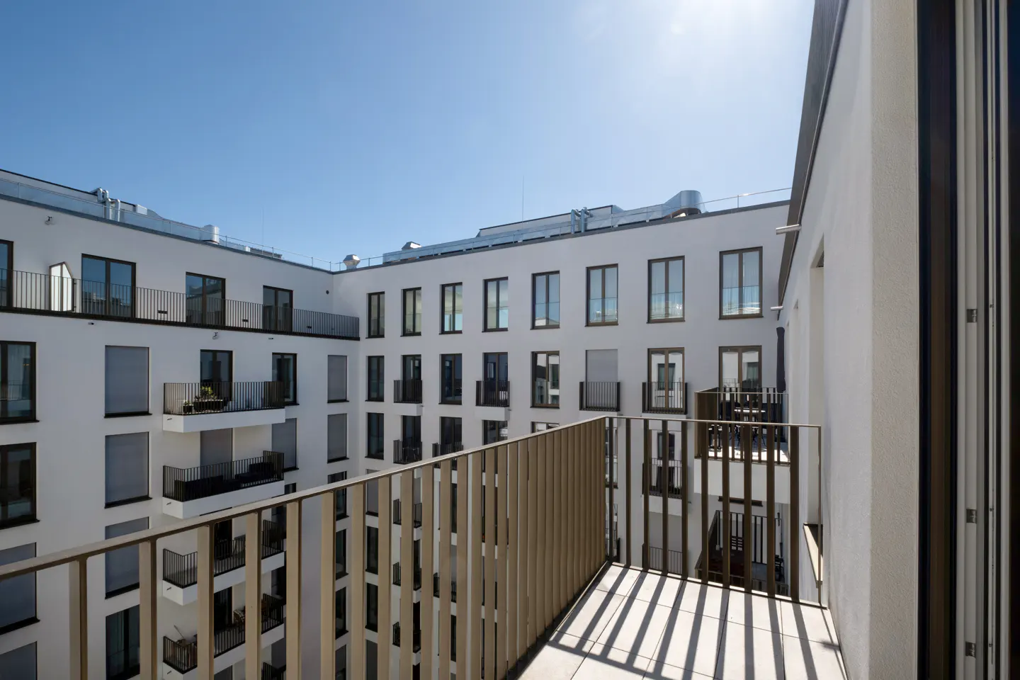 View from a balcony with a metal railing, overlooking a white apartment building with many windows and balconies under a clear blue sky.