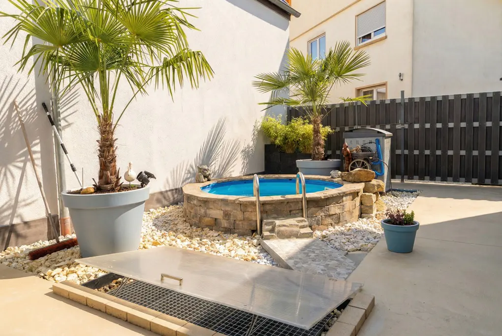 Backyard with a small, round, stone-walled pool filled with blue water, surrounded by white pebbles and potted palm trees.