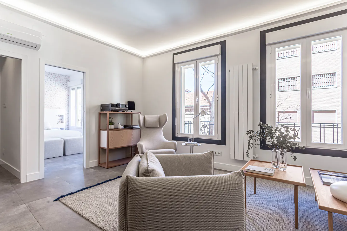 Bright living room with beige sofa, wingback chair, and wood shelving. Large windows with black trim offer a city view. A bedroom is visible through an open doorway.