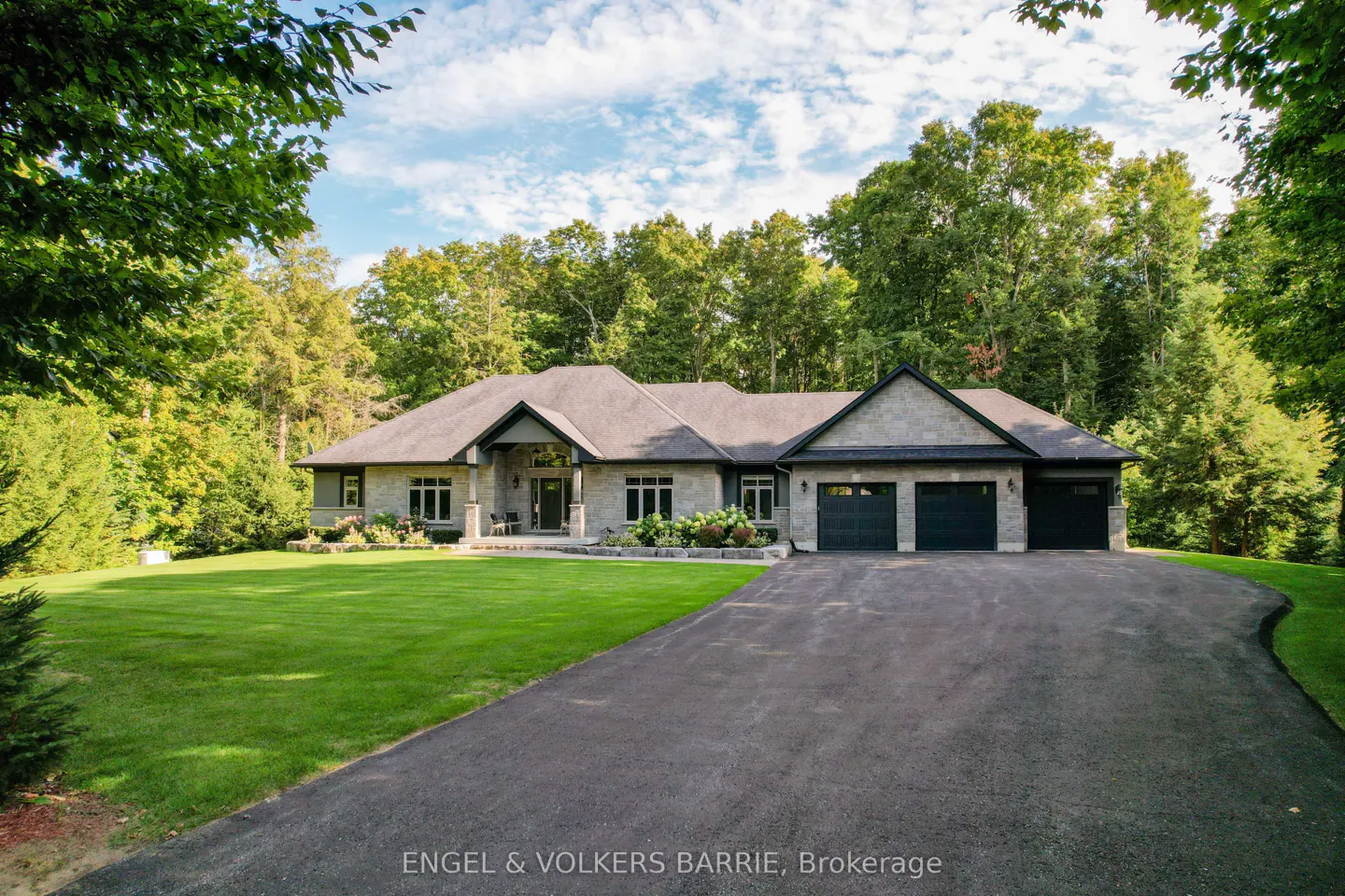 A single-story stone house with a dark gray roof and a three-car garage is surrounded by green trees and a lawn.