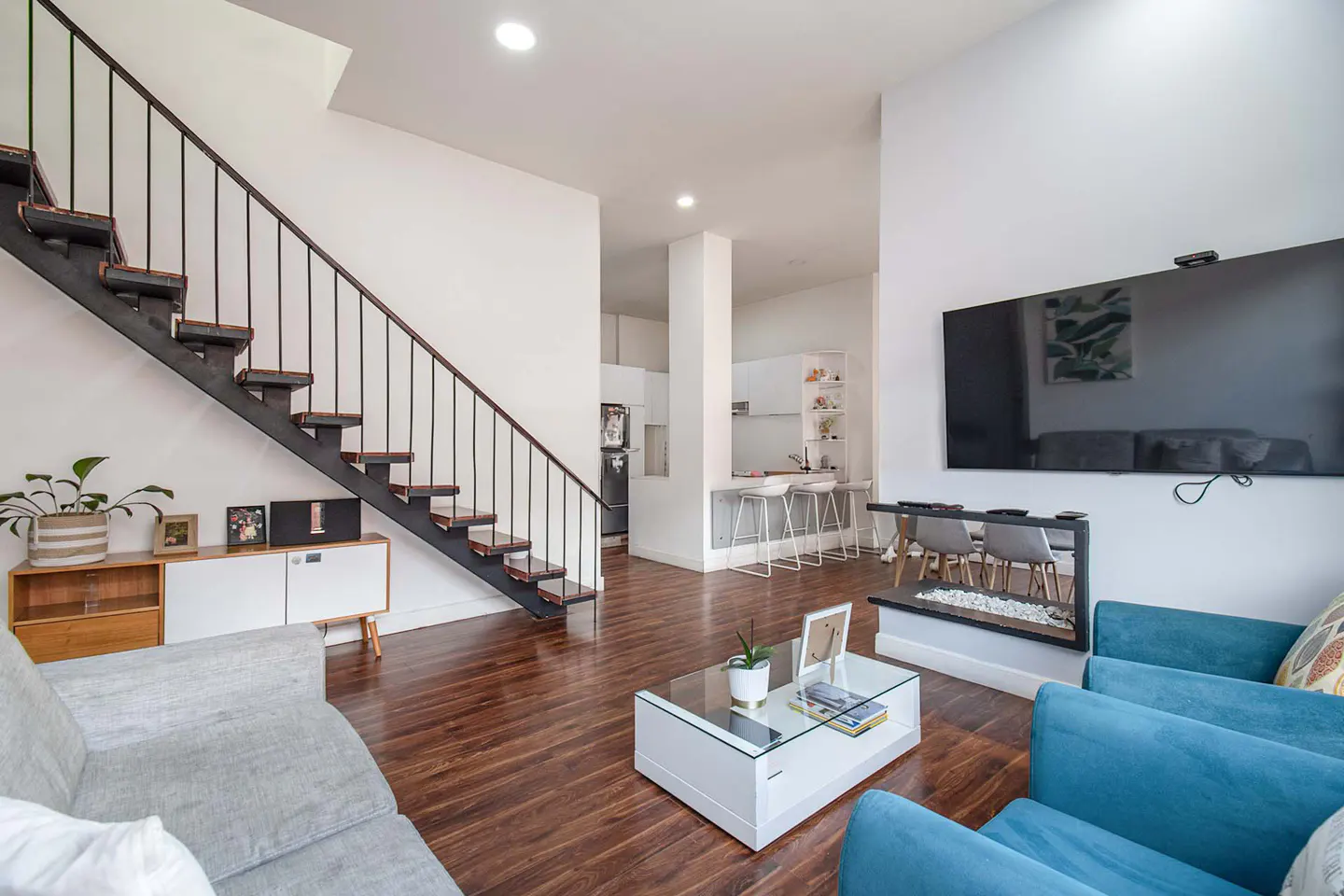 Bright living room with wood floors, white walls, and a black staircase. Blue armchairs face a TV above a modern fireplace. A white kitchen is visible in the background.