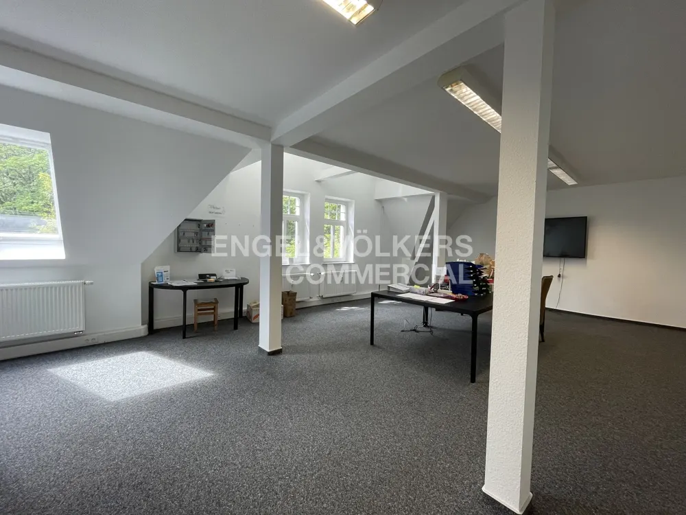 Bright, empty office space with white walls, gray carpet, and support columns. Desks and a TV are visible. Engel & Völkers logo in the center.