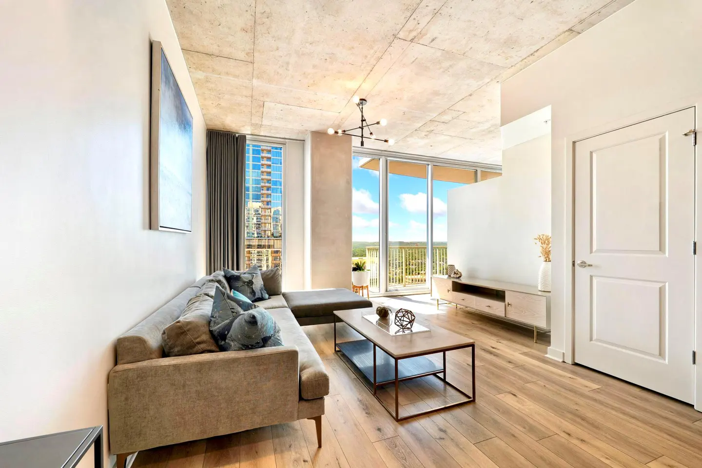 Modern living room with a grey sectional sofa, wood floors, concrete ceiling, and floor-to-ceiling windows with city views.