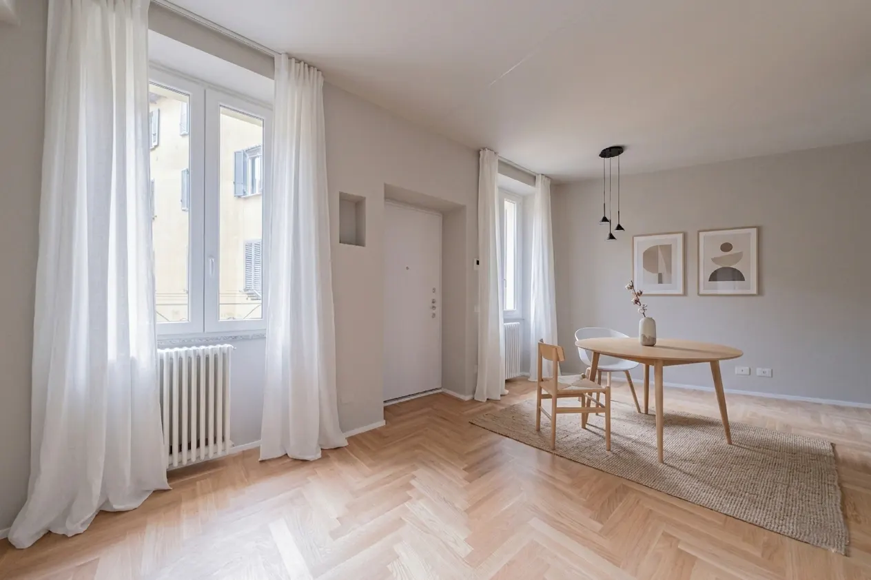 Bright, neutral dining room with wood floors, a round table, and white chairs. White curtains frame the windows. Two framed art prints hang on the wall.