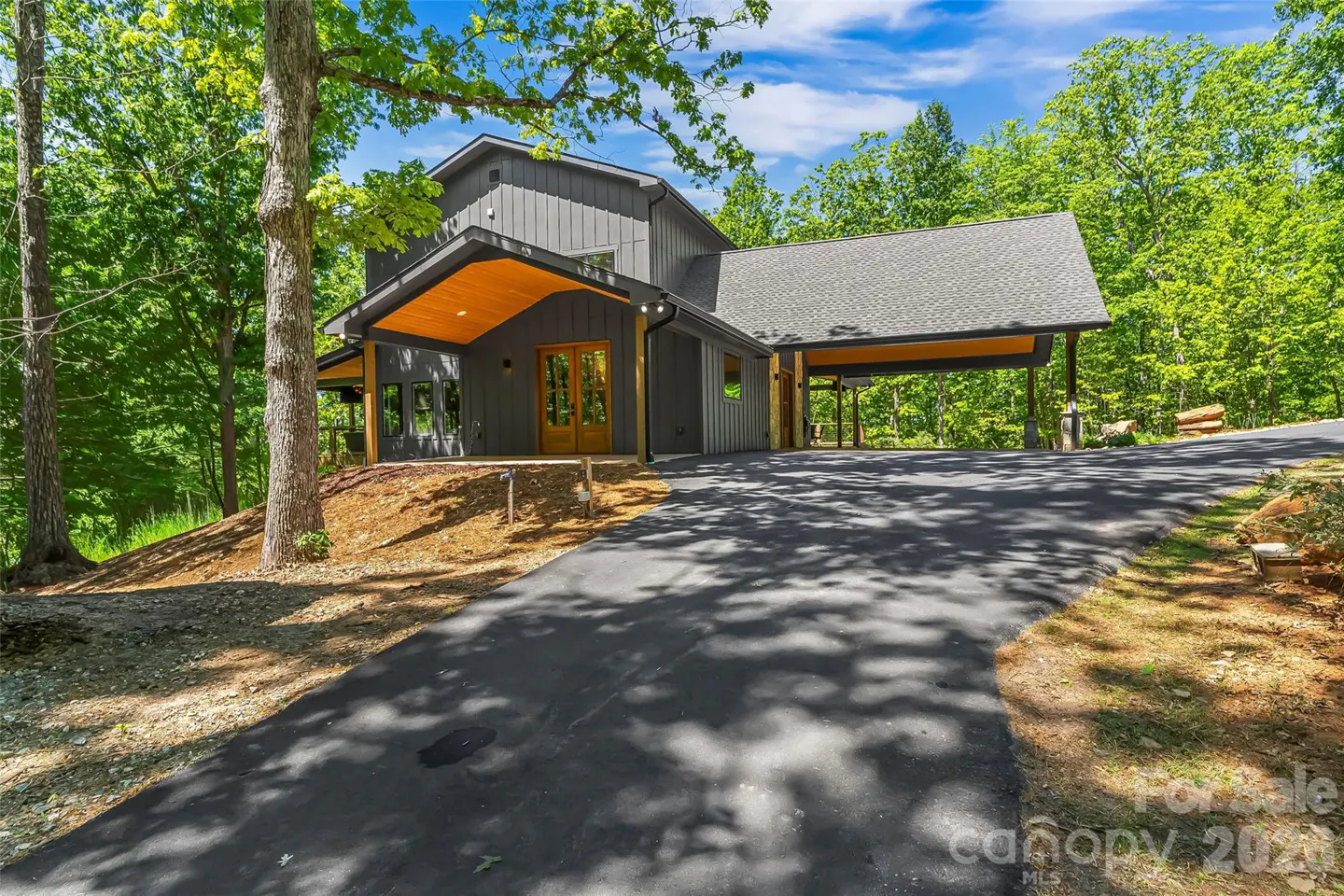 Modern gray house with wood accents, a covered driveway, and a long asphalt driveway surrounded by green trees.