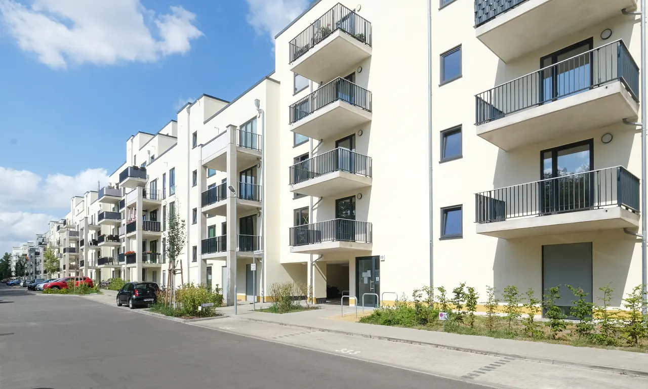Row of modern, white apartment buildings with black balconies under a blue sky. Cars parked along the street.