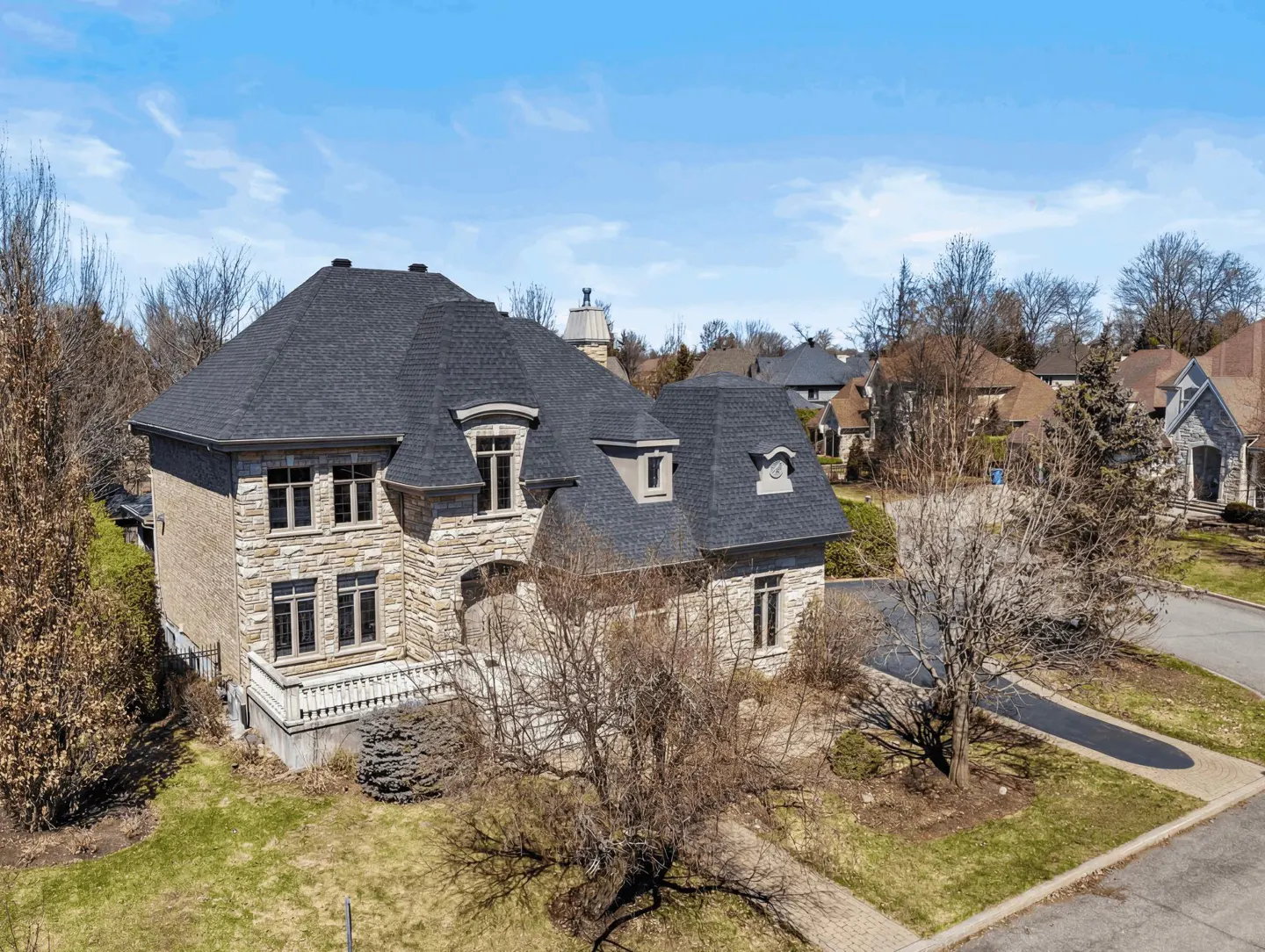 Aerial view of a large, two-story stone house with a dark gray roof, surrounded by trees and green lawns under a blue sky.
