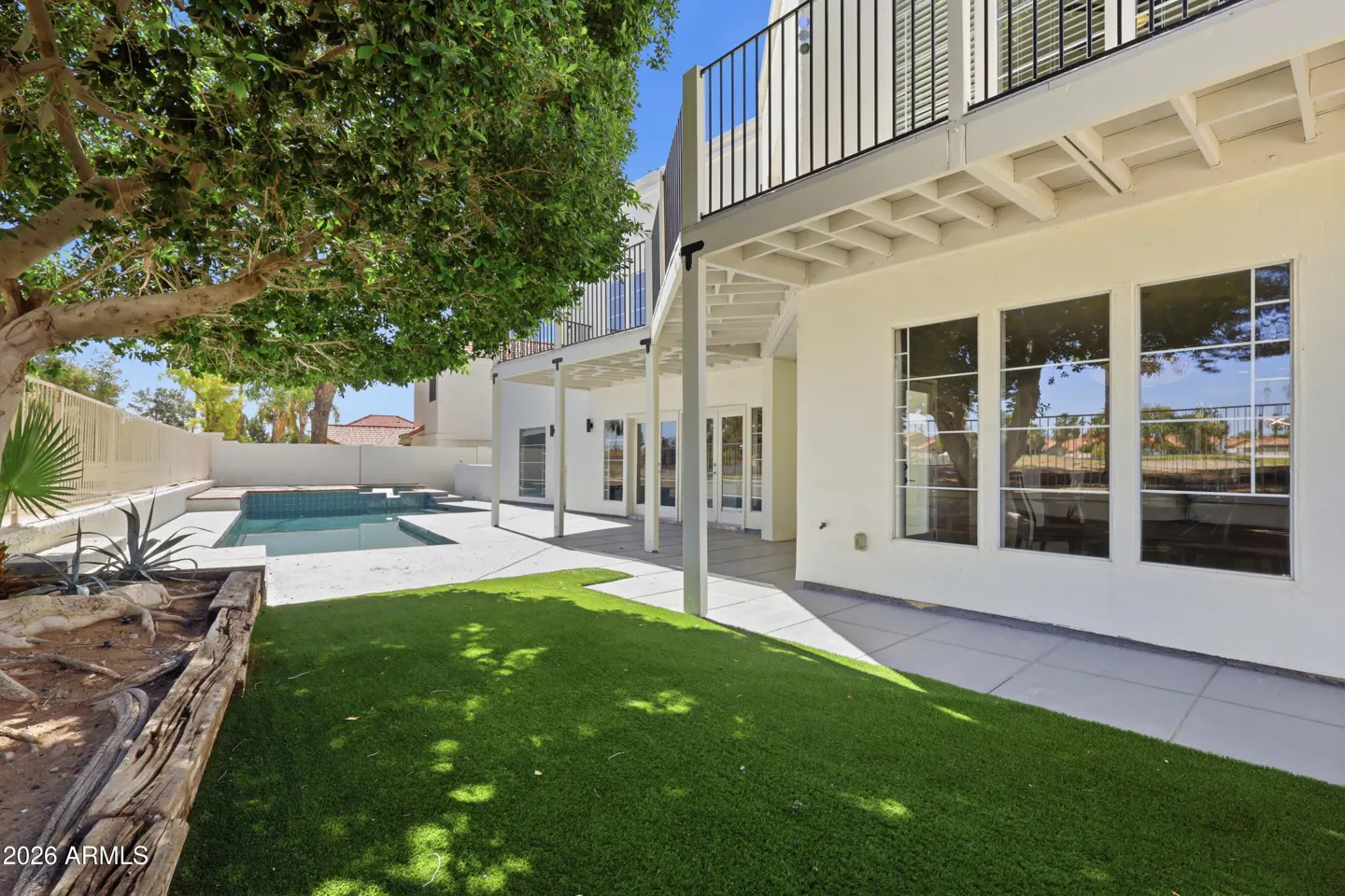 Backyard view of a two-story white house with a pool, green lawn, and a large tree. Balcony with black railings is visible.