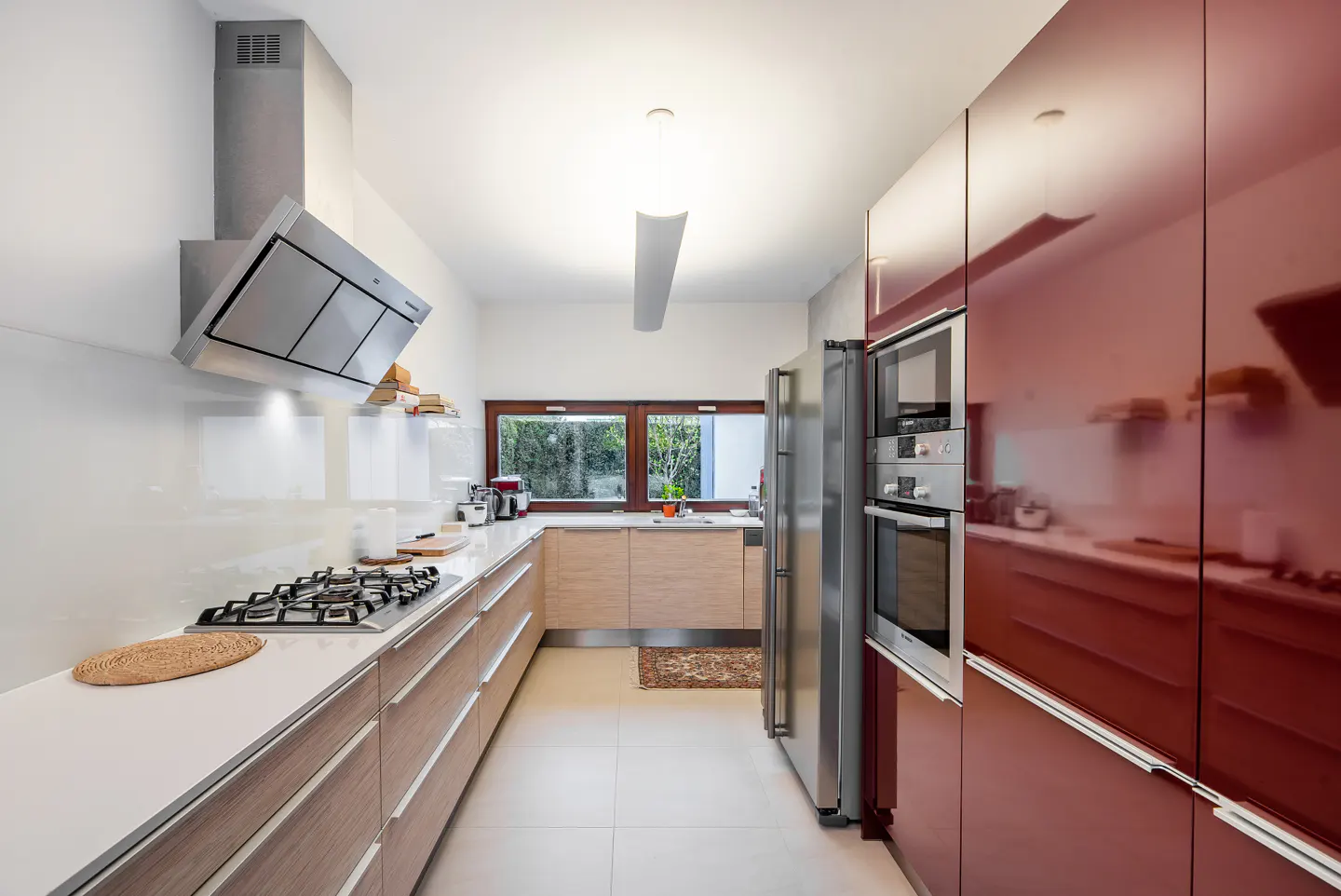 A modern kitchen with white walls, light wood cabinets, and red cabinets. Stainless steel appliances and a window are visible.