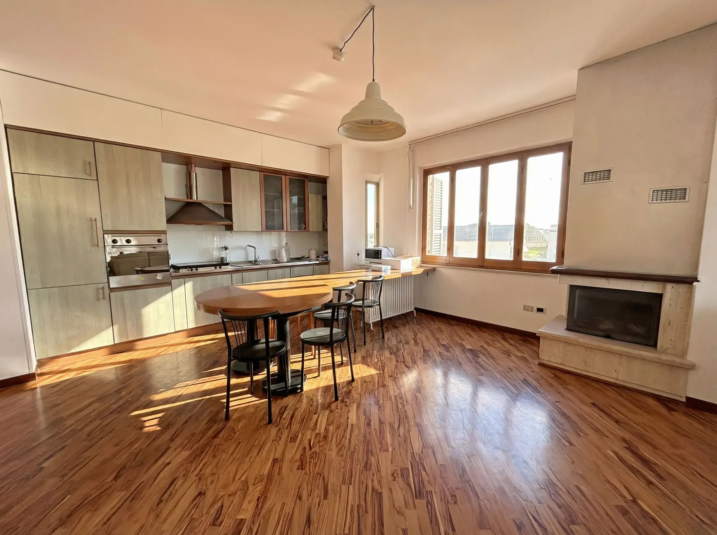 Open-concept kitchen and living room with wood floors, cabinets, round table with chairs, fireplace, and large window.