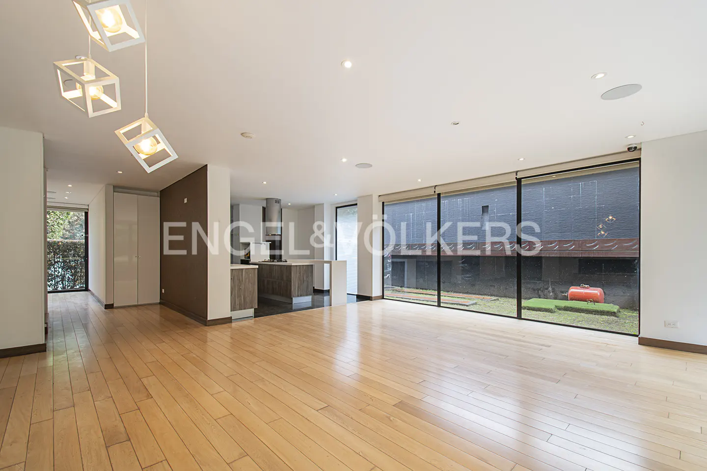Bright, open-plan living space with wood floors, modern lighting, and large windows overlooking a garden. Kitchen island visible in the background.