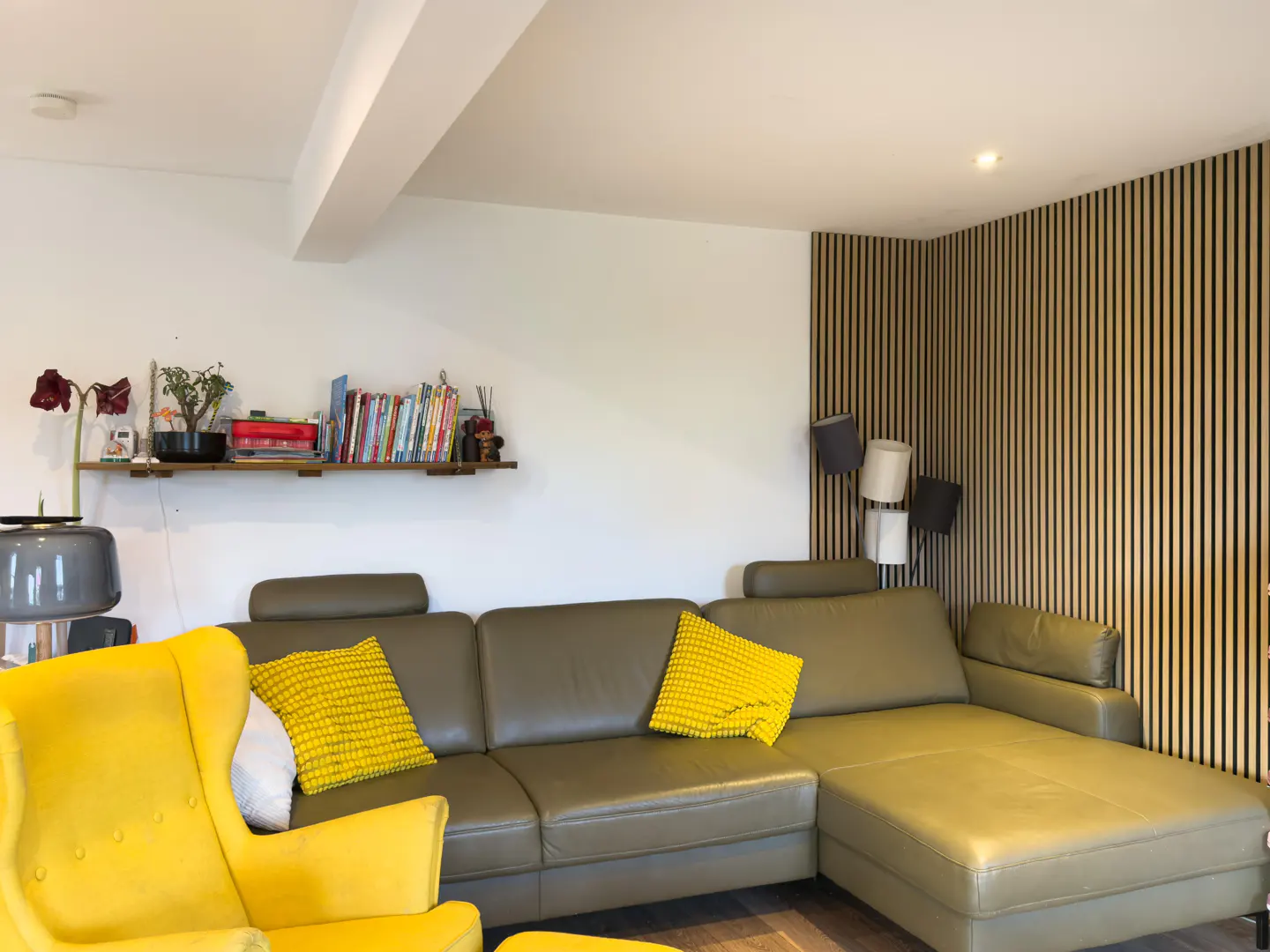 Living room with a green leather sectional sofa, yellow armchair, and wood slat wall. A shelf with books and plants is on the white wall.