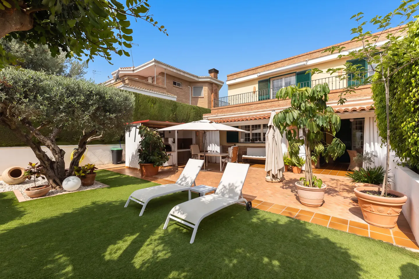 Sunny backyard patio with two white lounge chairs on a tiled terrace, artificial grass, potted plants, and a house exterior.